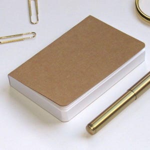 May include: A small, rectangular notebook with a brown cover and white pages. A gold pen, two gold paper clips, and gold scissors are arranged around the notebook on a white surface. Stationery supplies.