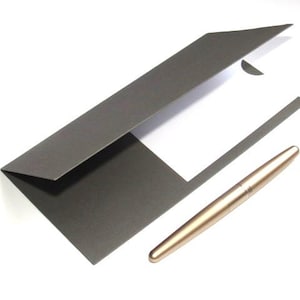 May include: A dark gray presentation folder with a white interior and a gold-colored pen. The pen is smooth and cylindrical. The folder is open, revealing a white sheet of paper.