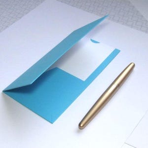 May include: A light blue paper folder with a white insert and a gold pen on a white surface. The folder is open, revealing the interior. The pen is lying next to the folder.