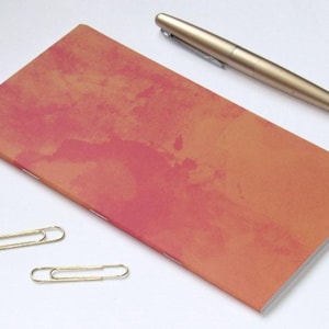 May include: A rectangular notebook with a mottled orange and pink cover, featuring a saddle-stitched spine. Beside the notebook lies a metallic gold fountain pen and two gold-toned metal paper clips, all arranged on a plain white surface.