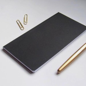 May include: A black notebook with a gold pen and two gold paper clips. The notebook is rectangular and has a textured cover. The pen is cylindrical and has a shiny finish. The paper clips are simple and elegant.