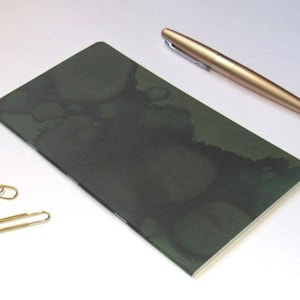 May include: A dark green notebook with a watercolor-like design, a gold pen, and two gold paper clips. The notebook is rectangular and has a soft, textured appearance. The pen is metallic and cylindrical.