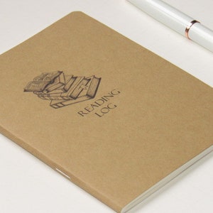 May include: A brown reading log notebook with the words "READING LOG" printed on the cover. The cover also features a graphic of stacked books. A white pen with rose gold accents rests on the upper right corner.
