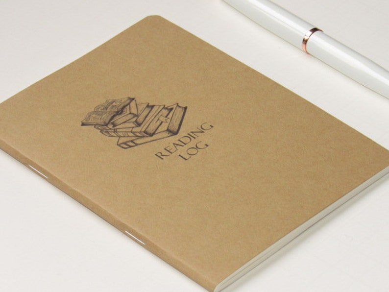 May include: A brown reading log notebook with the words "READING LOG" printed on the cover. The cover also features a graphic of stacked books. A white pen with rose gold accents rests on the upper right corner.
