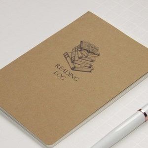 May include: A brown Reading Log notebook with a stack of books illustration and the text "Reading Log". A white pen with rose gold accents rests on the surface next to the notebook.