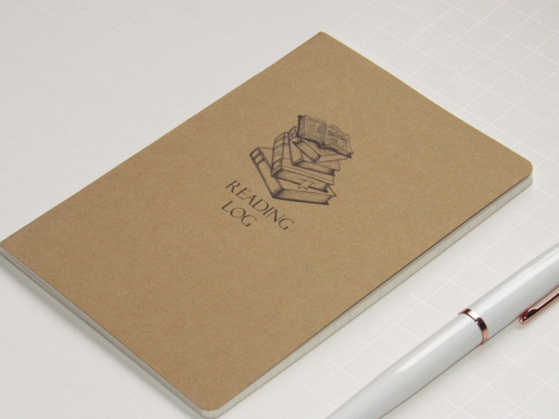 May include: A brown Reading Log notebook with a stack of books illustration and the text "Reading Log". A white pen with rose gold accents rests on the surface next to the notebook.
