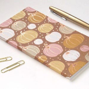 May include: A rectangular notebook with a brown cover featuring a repeating pattern of pumpkins in shades of gold, white, and soft pink, accented by small white dots. A gold-toned pen and two gold-colored metal paper clips are placed nearby.