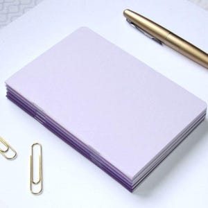 May include: A stack of lavender-colored notepaper with purple edges, a gold pen, and two gold paper clips on a white surface. The paper has rounded corners. The pen is lying diagonally across the top of the paper.