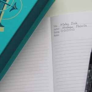 May include: An open notebook with the title "Moby Dick" and author Herman Melville, with a rating of three stars. A black pen rests on the lined pages. A teal book with gold accents is in the background.