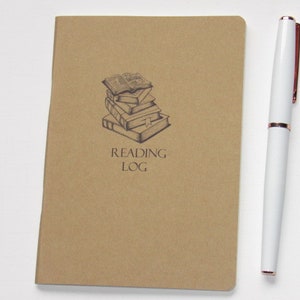 May include: A tan Reading Log journal with a stack of books illustration and the words "Reading Log" printed below. A white pen with rose gold accents rests beside the journal.