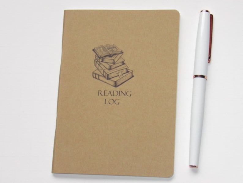 May include: A tan Reading Log journal with a stack of books illustration and the words "Reading Log" printed below. A white pen with rose gold accents rests beside the journal.