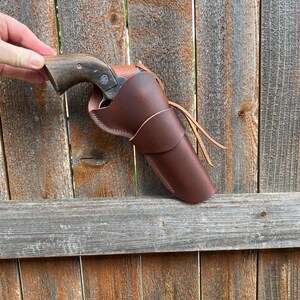 May include: A brown leather holster with a revolver. The holster is a rich, dark brown and features a stitched design. The revolver has a wooden handle and a silver-colored barrel. The image is set against a weathered wooden fence.