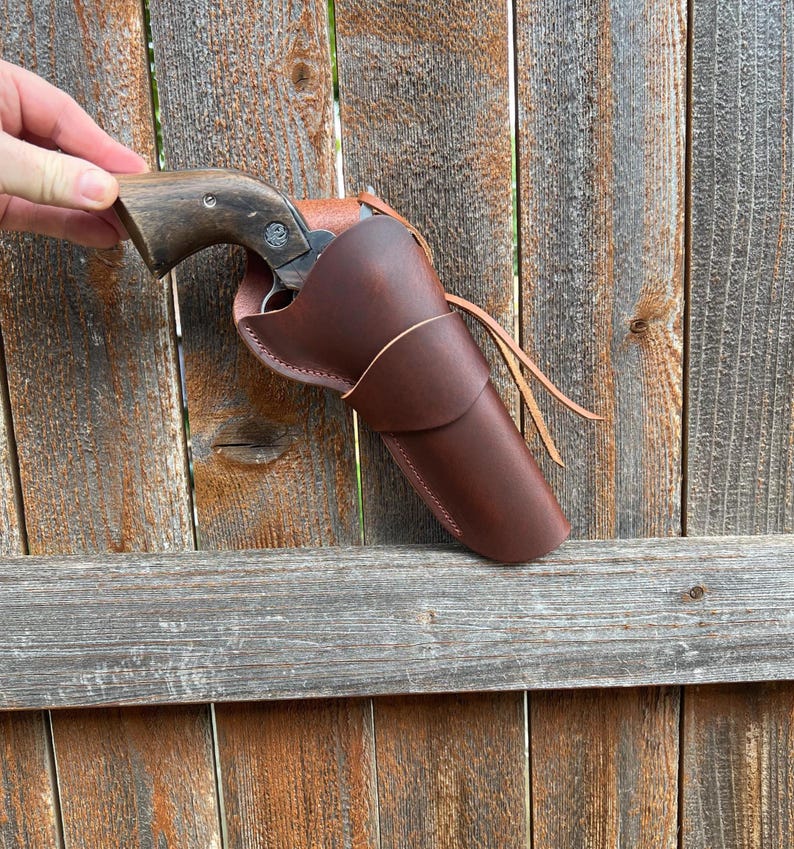 May include: A brown leather holster with a revolver. The holster is a rich, dark brown and features a stitched design. The revolver has a wooden handle and a silver-colored barrel. The image is set against a weathered wooden fence.