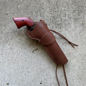 May include: A brown leather holster with a matching brown revolver. The revolver has a dark wood handle. The holster is secured with a leather tie. The items are resting on a gray concrete surface.