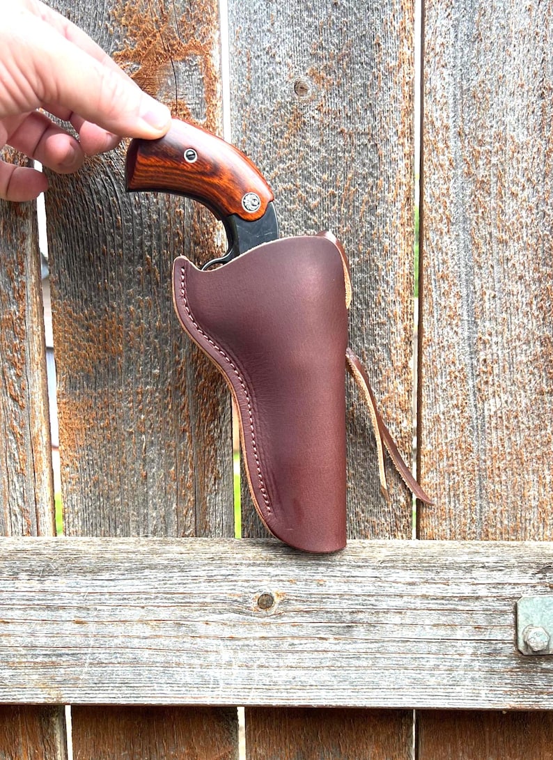 May include: A brown leather holster with a stitched edge holds a knife with a wooden handle. The knife is partially visible. The background is a weathered wooden fence.