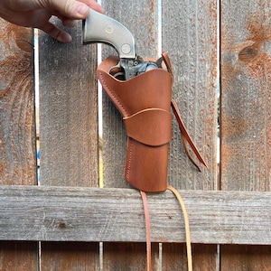 May include: A silver revolver in a brown leather holster. The holster has a stitched design and long leather straps. The revolver is held against a weathered wooden fence.