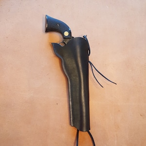 May include: A black leather holster with a brown strap and a black gun inside. The holster is hanging on a brown leather surface.