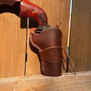 May include: A vintage revolver with a dark wood grip is partially inserted into a brown leather holster. The holster is secured with a leather strap. The background is a wooden fence.