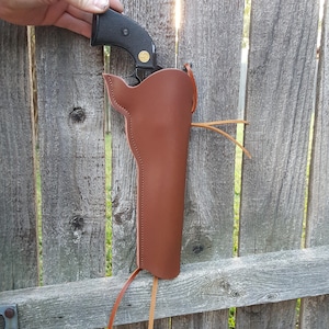May include: A brown leather holster with a black handgun. The holster is designed to be worn on the hip and has a stitched design. The handgun has a black grip and a gold emblem. The holster is hanging on a wooden fence.