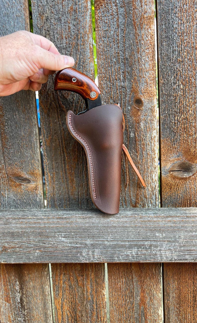 K&ouml;nnte beinhalten: Eine braune Lederholster mit einem Messer. Das Messer hat einen Holzgriff mit dunklem Finish. Die Holster ist mit hellfarbenem Faden gen&auml;ht und hat einen Lederriemen.