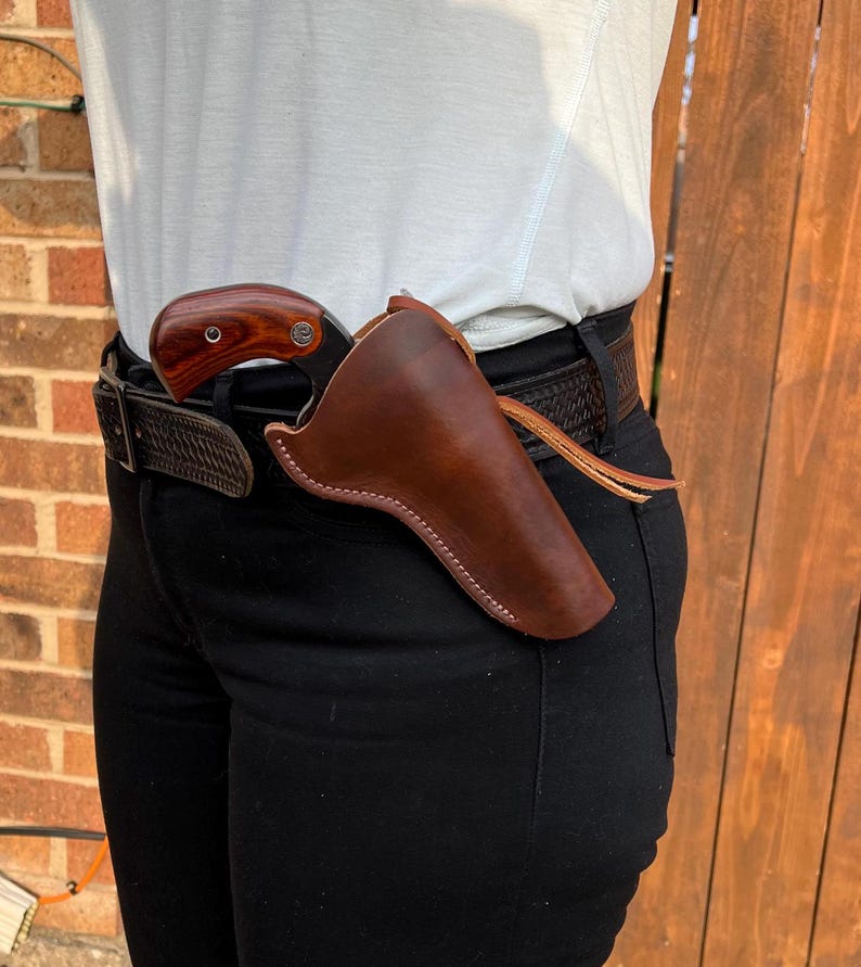 Op de afbeelding: Een bruine leren holster met een pistool erin. De holster is bevestigd aan een zwarte riem.