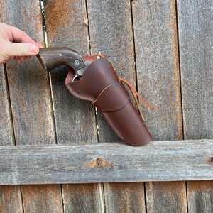May include: A brown leather holster with a revolver inside. The holster is a rich, dark brown color and features a simple, classic design. The revolver has a wooden grip and a silver barrel. The holster is held against a weathered wooden fence.