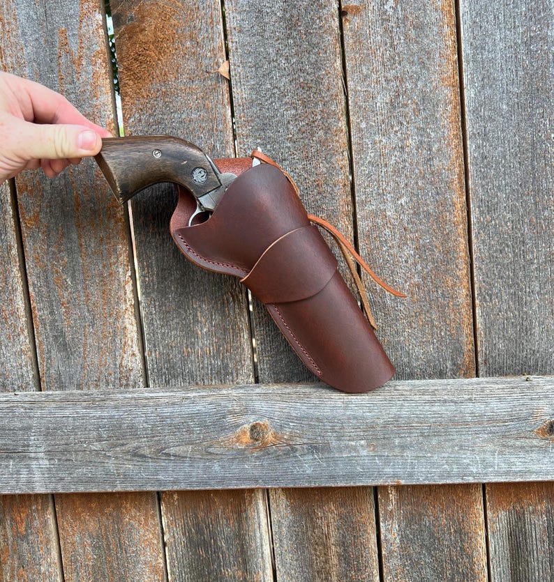 May include: A brown leather holster with a revolver inside. The holster is a rich, dark brown color and features a simple, classic design. The revolver has a wooden grip and a silver barrel. The holster is held against a weathered wooden fence.