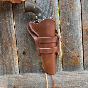 May include: A brown leather holster with a matching revolver. The holster has two belt loops and a long strap. The revolver has a dark wooden handle and a silver barrel. The items are displayed against a weathered wooden fence.