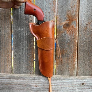 May include: A brown leather holster with a matching leather strap, holding a revolver with a dark wood handle. The holster is shown against a weathered wooden fence.