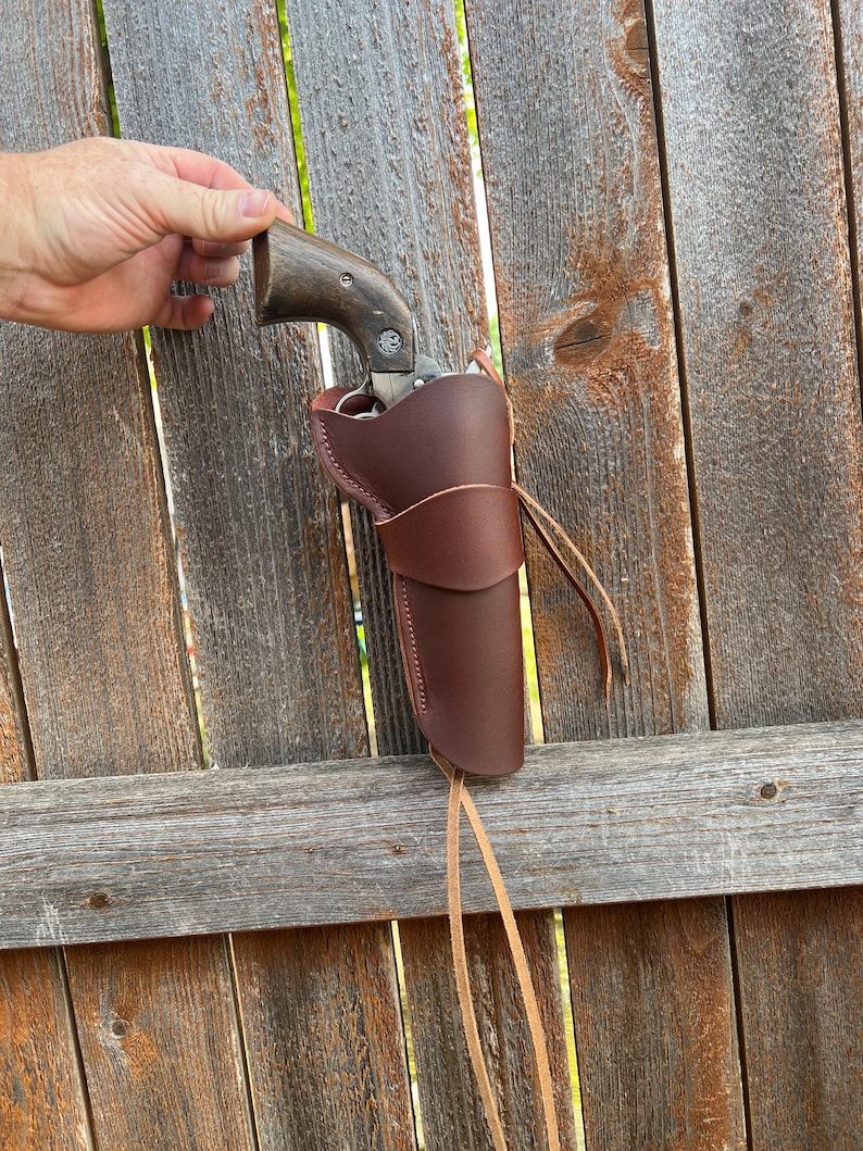 May include: A brown leather holster with a revolver. The holster is designed to hold a handgun and is secured with a leather tie. The revolver has a wooden handle. The background is a weathered wooden fence.