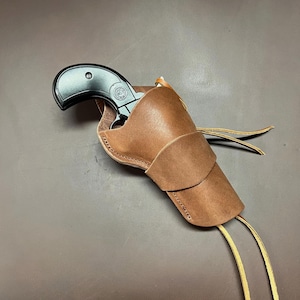 May include: A black handgun in a brown leather holster. The holster is made of stitched leather with a flap and two long, thin leather ties. The handgun is partially visible, with a black handle and a silver emblem.
