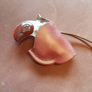 May include: A brown leather holster with a silver handgun inside. The holster has a flap that covers the gun and a strap that goes around the wrist.