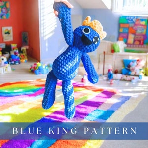 May include: A blue crocheted plush toy with a yellow crown and a big smile. The toy is being held up in the air against a colorful rainbow striped rug. The text "BLUE KING PATTERN" is at the bottom of the image.