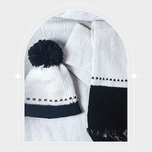 Op de afbeelding: Een gebreide wintermuts met een zwarte pompon en een bijpassende sjaal. De muts en sjaal zijn wit met een zwarte rand en decoratieve stiksels. De sjaal heeft een gefranjerde rand.