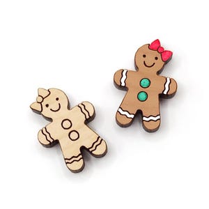 May include: Two wooden gingerbread people ornaments. One is brown with green buttons and a red bow. The other is natural wood with black outlines and no paint.