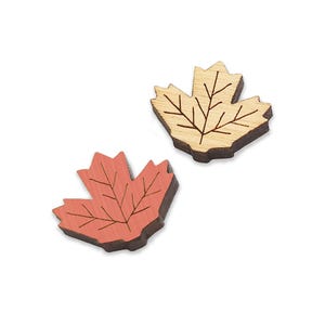 May include: Two wooden maple leaves, one is natural wood color and the other is painted a light coral color.