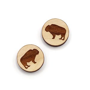 May include: Two round wooden earrings with a light wood grain. Each earring features a dark brown bison silhouette. The earrings are set against a white background.