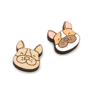 May include: Two wooden French bulldog head-shaped embellishments. One is unpainted with etched details, the other is painted with brown, tan, white, and pink accents. The dog faces have a smiling expression and are set against a white background.