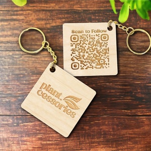 May include: Two wooden keychains with gold-coloured key rings. One keychain has the text "plant.cessories" and a leaf design. The other keychain has a QR code and the text "Scan to Follow". The keychains are on a dark wood surface.