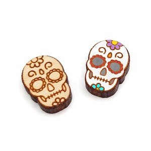 May include: Two wooden sugar skull charms with colorful painted details. One skull is white with red and blue accents, the other is natural wood with brown accents.