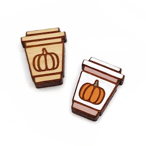 May include: Two wooden coffee cup ornaments with a pumpkin design. One cup is brown and the other is white with a pink rim.