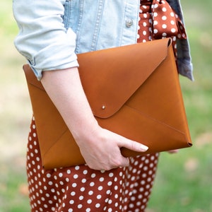 May include: A brown leather envelope clutch with a flap closure. The clutch is held by a person wearing a polka dot dress and a denim jacket.