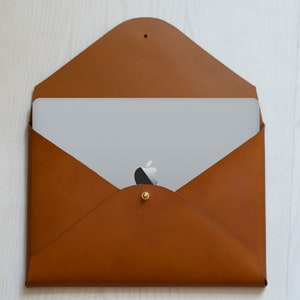 Puede incluir: Una funda para port&aacute;til de cuero marr&oacute;n estilo sobre con cierre de bot&oacute;n dorado. Un port&aacute;til est&aacute; dentro de la funda, con el logotipo de Apple visible.