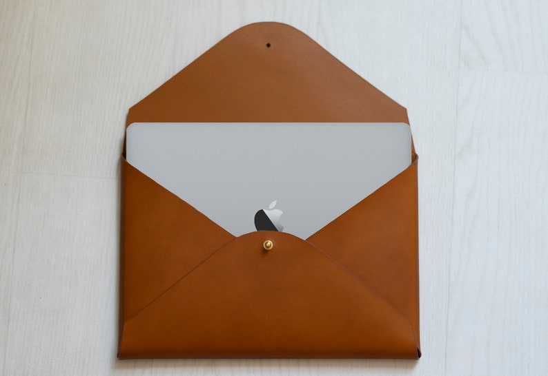 Puede incluir: Una funda para port&aacute;til de cuero marr&oacute;n estilo sobre con cierre de bot&oacute;n dorado. Un port&aacute;til est&aacute; dentro de la funda, con el logotipo de Apple visible.
