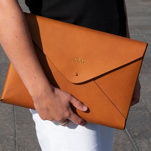 Puede incluir: Un clutch de cuero marr&oacute;n en forma de sobre con un monograma "CLG" en relieve dorado.
