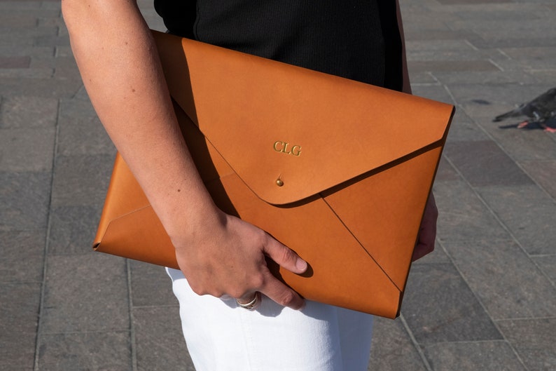 Puede incluir: Un clutch de cuero marr&oacute;n en forma de sobre con un monograma "CLG" en relieve dorado.