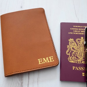 May include: A brown leather passport holder with the initials "EME" embossed in gold. The holder is next to a burgundy passport with a gold royal crest and the word "PASSPORT" printed on the cover.