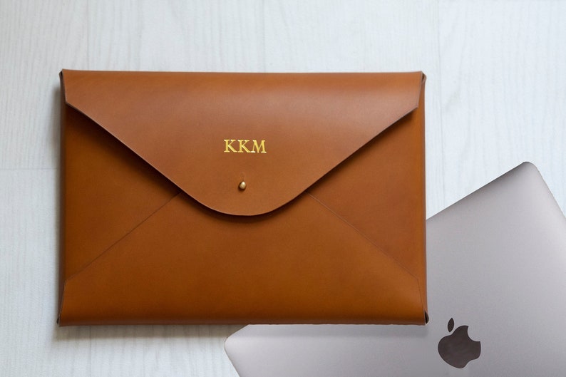 Puede incluir: Una funda para port&aacute;til de cuero marr&oacute;n con un monograma dorado "KKM" en la parte delantera.