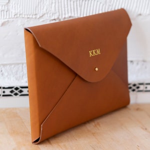 Puede incluir: Un clutch de cuero marr&oacute;n en forma de sobre con un monograma dorado "KKM" en la parte delantera.