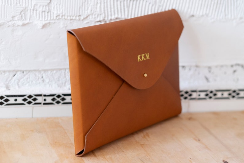 Puede incluir: Un clutch de cuero marr&oacute;n en forma de sobre con un monograma dorado "KKM" en la parte delantera.